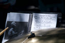 A spiral-bound jazz sheet music book open on a stand, showing musical notation, a photo of a man playing guitar, and part of a drum set is in the foreground.